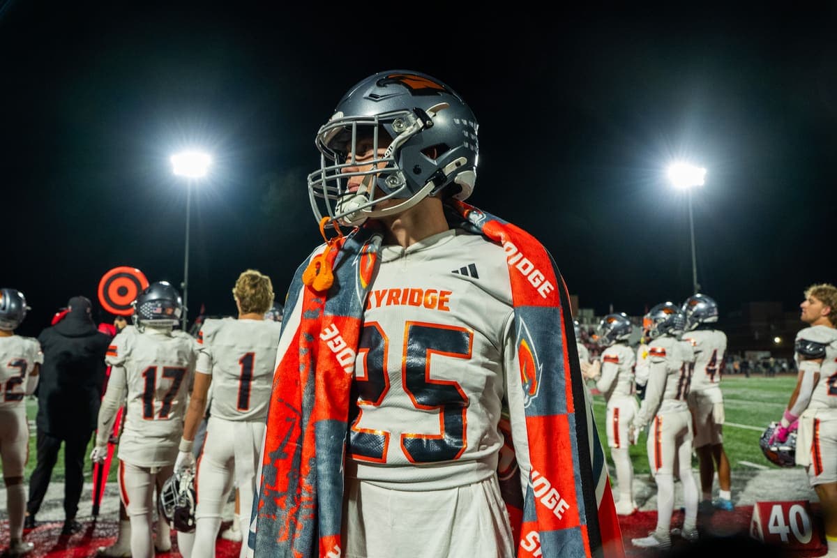 A Sky Ridge football player wearing his custom mascot blanket like a cape on the sideline under stadium lights